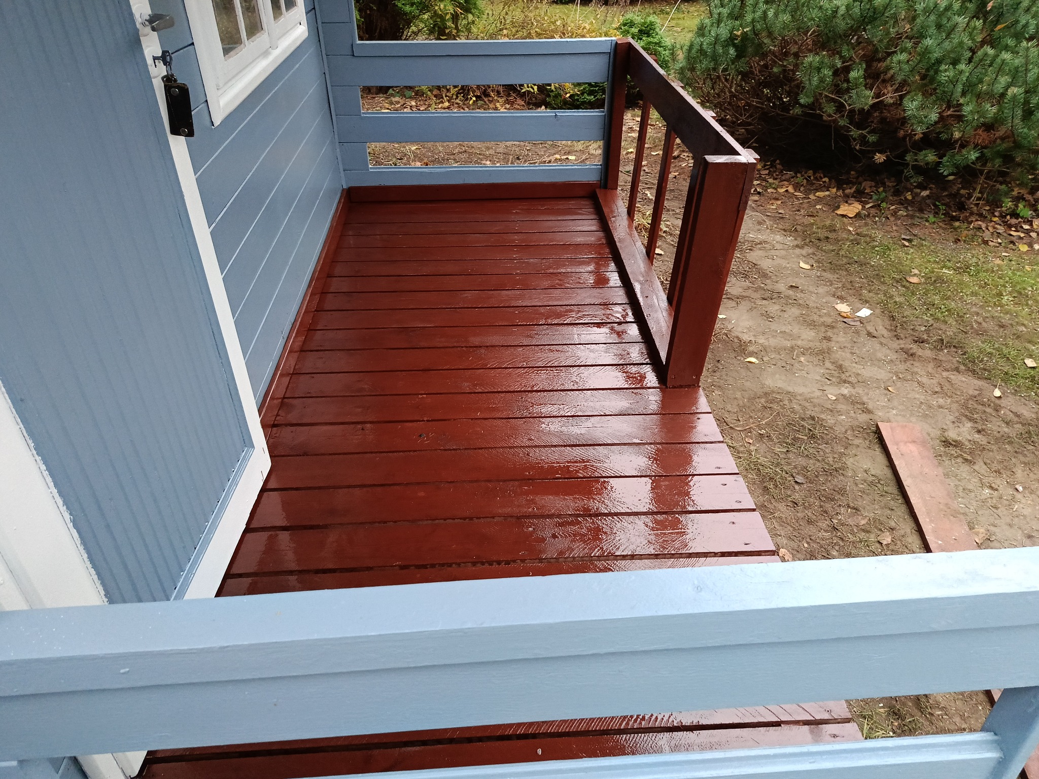 Świeżo pomalowany na brązowo drewniany taras przy domku z niebieskimi ścianami i białymi ramami okiennymi, widok z góry.