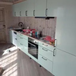 Montaż jasnej, nowoczesnej kuchni z drewnianym wykończeniem ścian i sufitu. Widoczne narzędzia i elementy montażowe w trakcie instalacji.