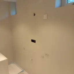 Gładka, beżowa ściana po szpachlowaniu z widocznymi puszkami elektrycznymi i gniazdami. Podłoga zabezpieczona folią malarską. Widok na okno.