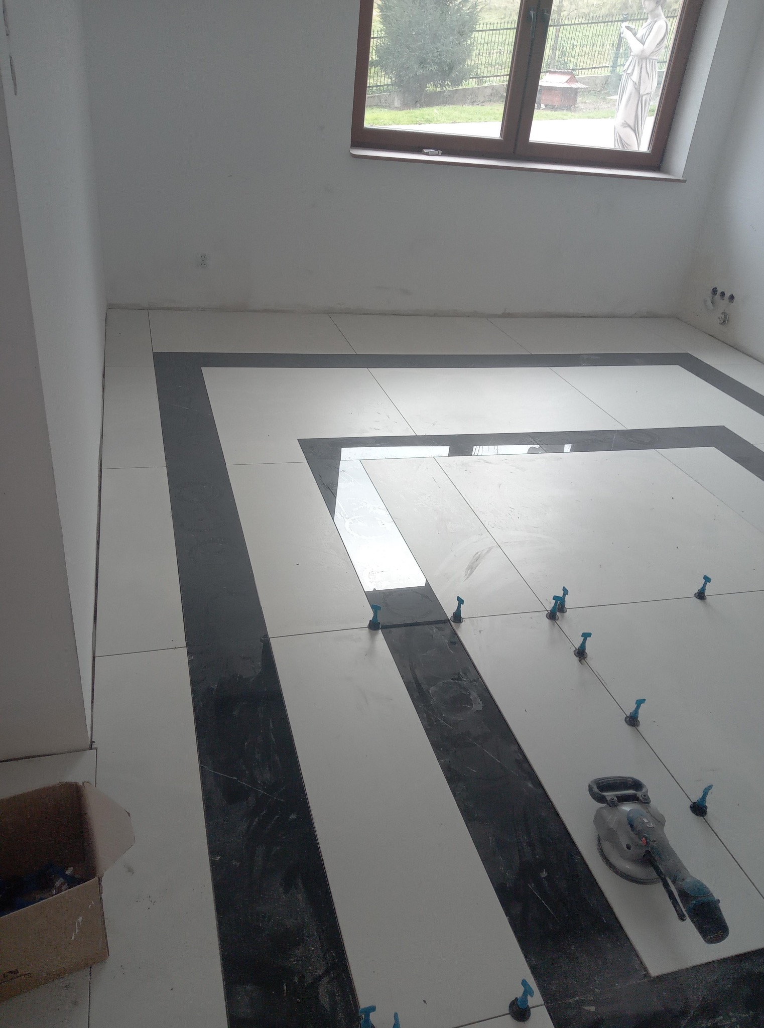 Układanie podłogi z płytek w geometryczny wzór, z użyciem narzędzi i poziomowania, w jasnym pomieszczeniu z oknem i widokiem na zewnątrz.