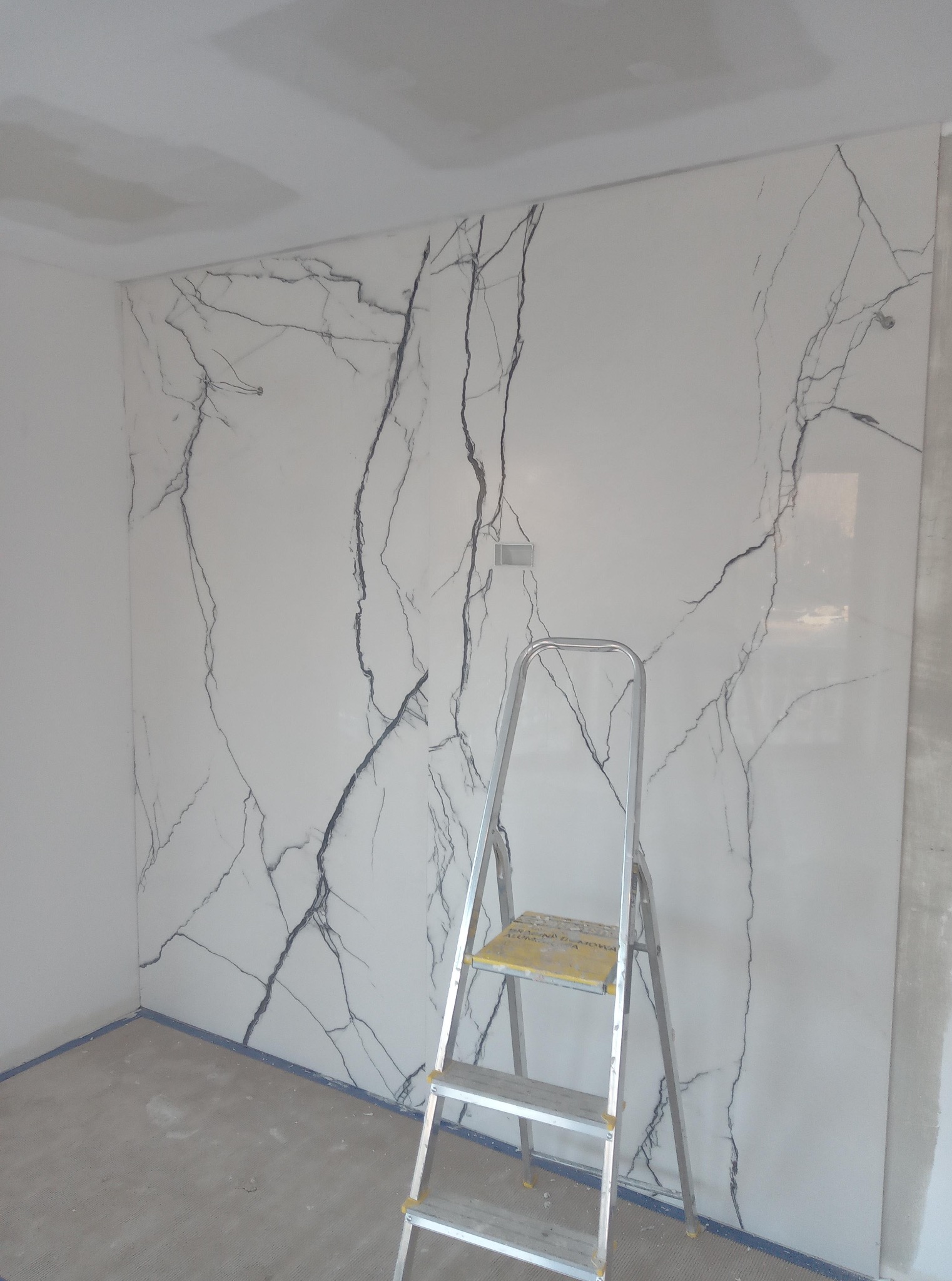 Ściana wykończona płytami imitującymi marmur, z widoczną drabiną, sugerująca prace wykończeniowe w trakcie realizacji. Estetyczne wnętrze, nowoczesny design.