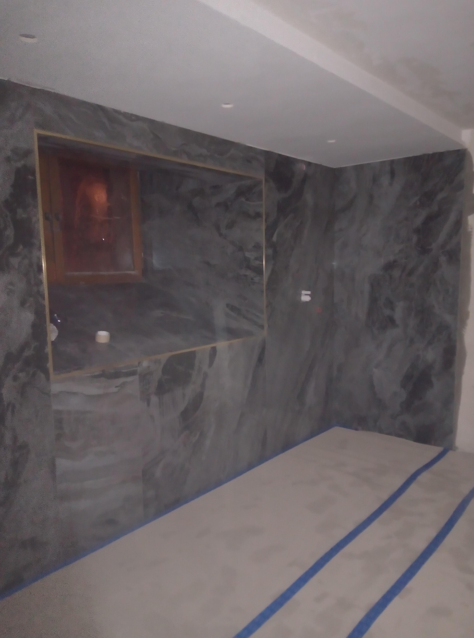 Wykończona łazienka z szarymi płytkami imitującymi marmur i złotymi detalami. Podłoga zabezpieczona taśmą malarską. Widoczny fragment lustra.