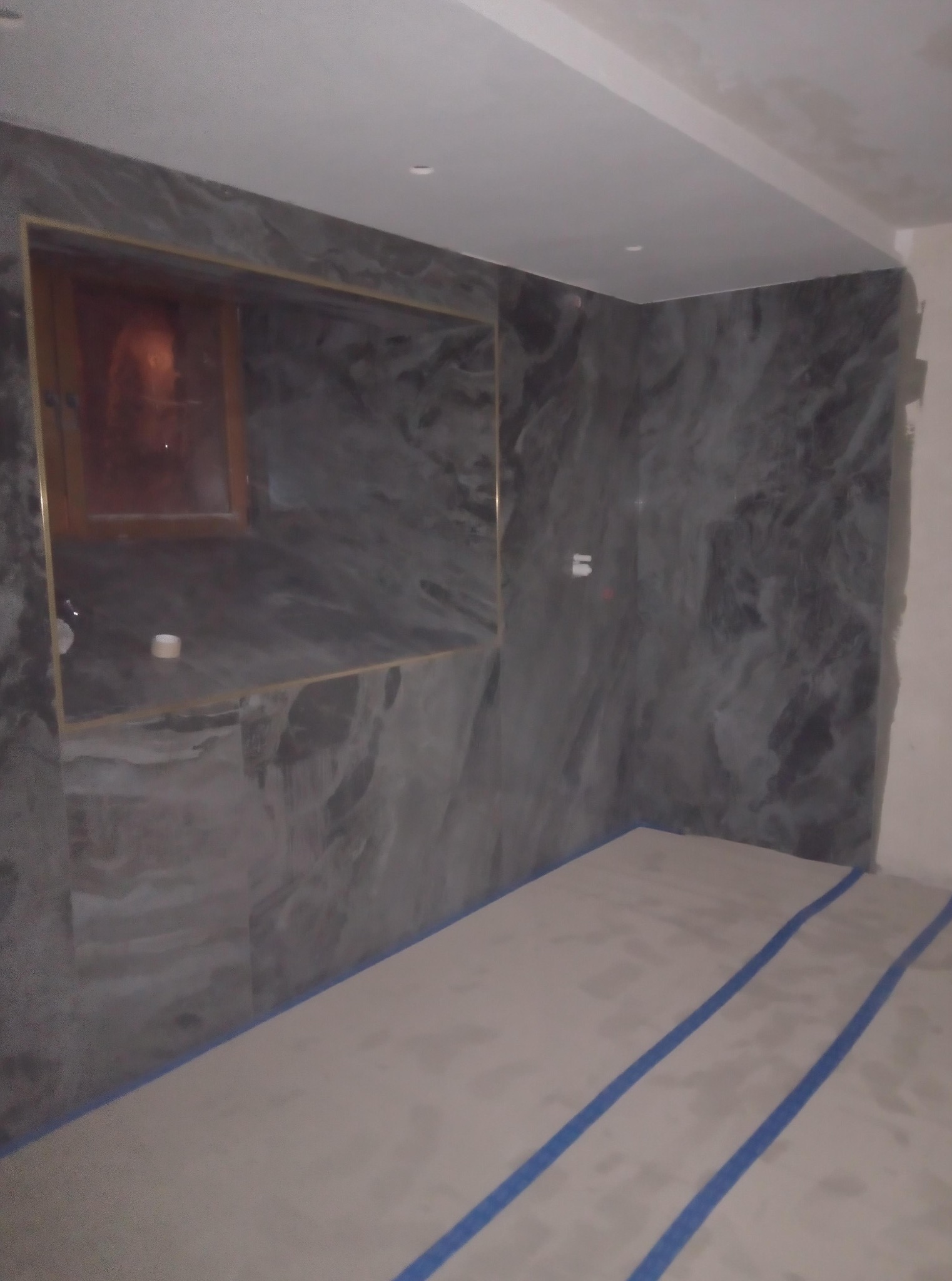 Wykończona łazienka z szarymi płytkami imitującymi marmur na ścianach, widoczne zabezpieczenie podłogi taśmą malarską. Wnęka z oświetleniem.