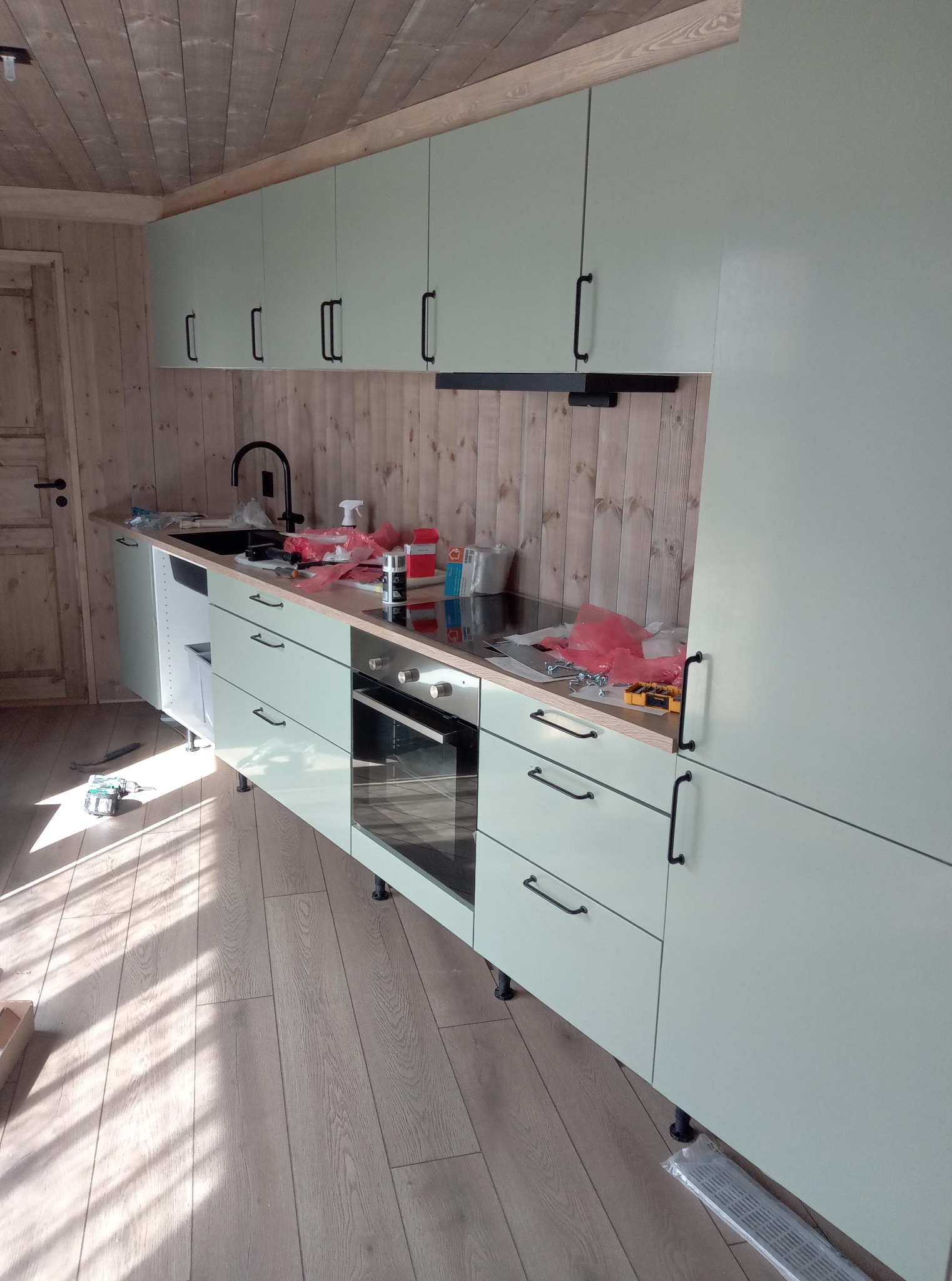 Montaż jasnej, nowoczesnej kuchni z drewnianym wykończeniem ścian i sufitu. Widoczne narzędzia i elementy montażowe w trakcie instalacji.