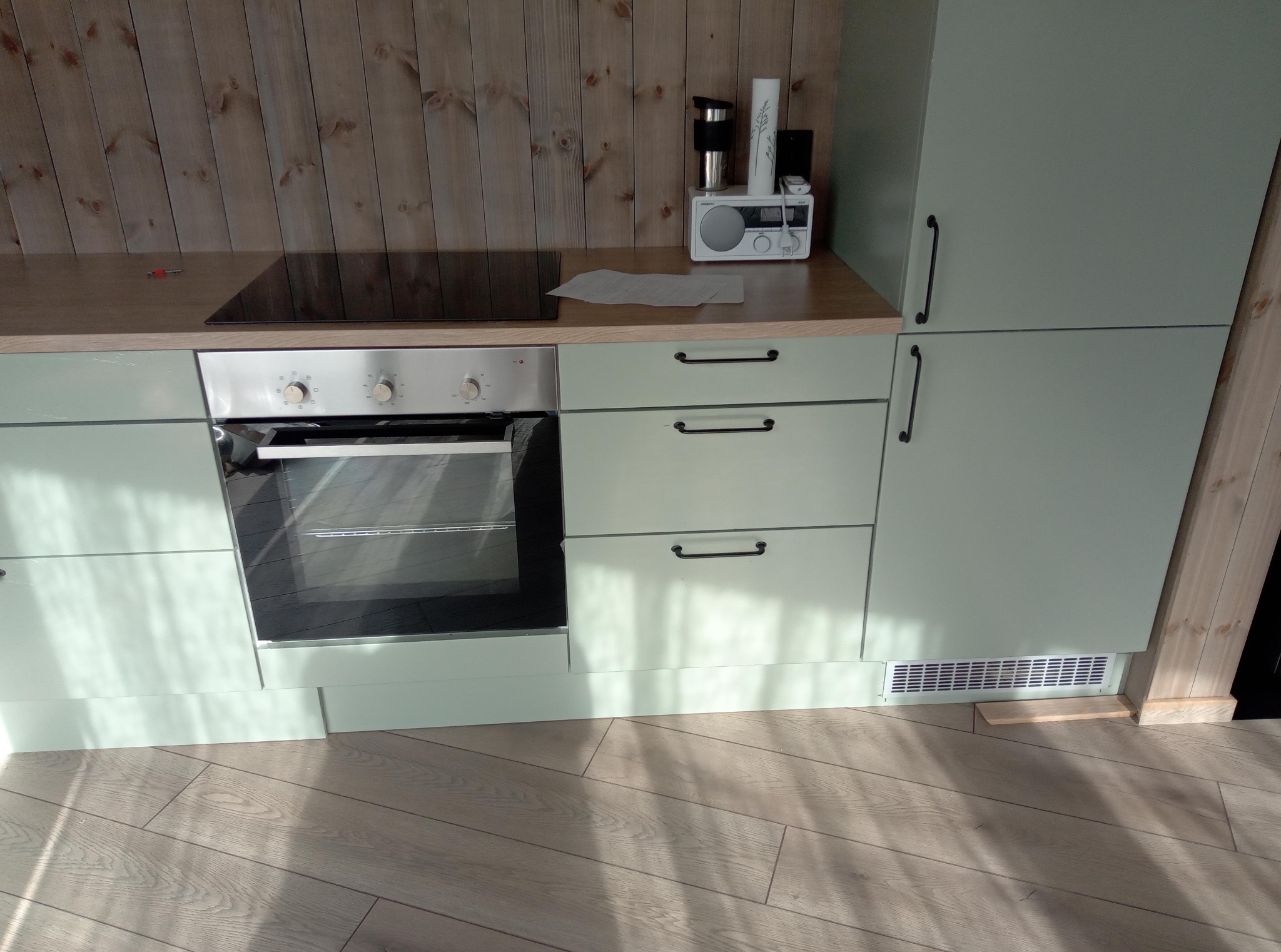 Jasnozielona zabudowa kuchenna z drewnianym blatem i srebrnym piekarnikiem na tle drewnianej ściany. Widoczny fragment podłogi z paneli.