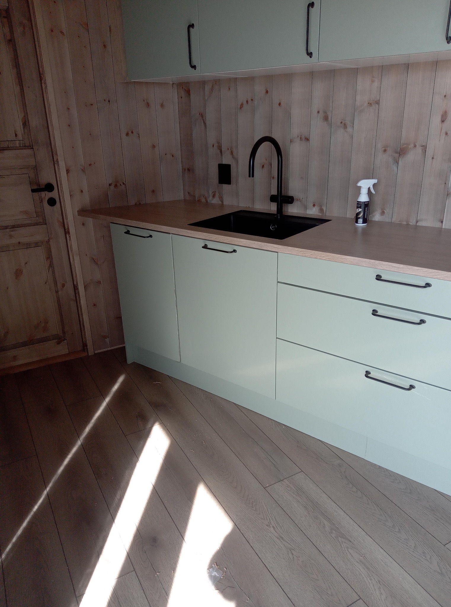 Jasna, drewniana kuchnia z czarną armaturą i subtelnymi, zielonymi szafkami. Ujęcie z niskiego kąta, podkreślające drewniane wykończenie ścian i podłogi.