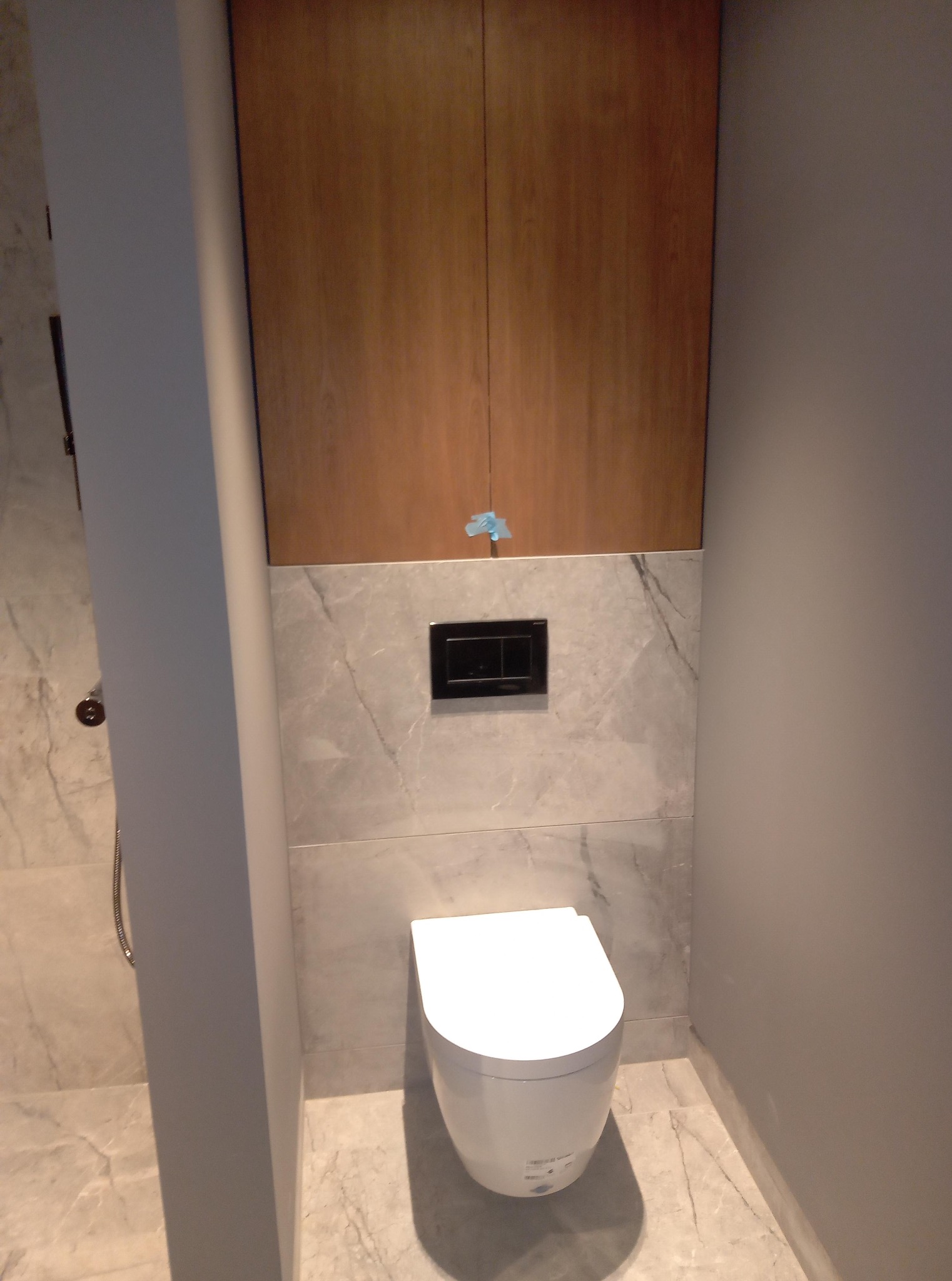 Nowoczesna toaleta z zabudową z drewna i marmurowymi płytkami. Minimalistyczny design, wisząca miska WC, czarny panel sterujący. Wykończenie łazienki w neutralnych barwach.