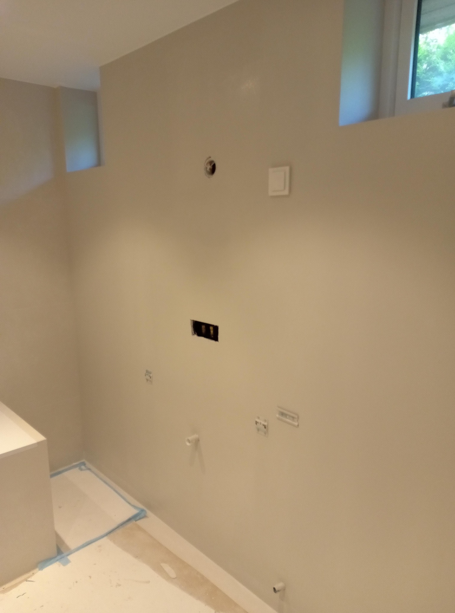 Gładka, beżowa ściana po szpachlowaniu z widocznymi puszkami elektrycznymi i gniazdami. Podłoga zabezpieczona folią malarską. Widok na okno.