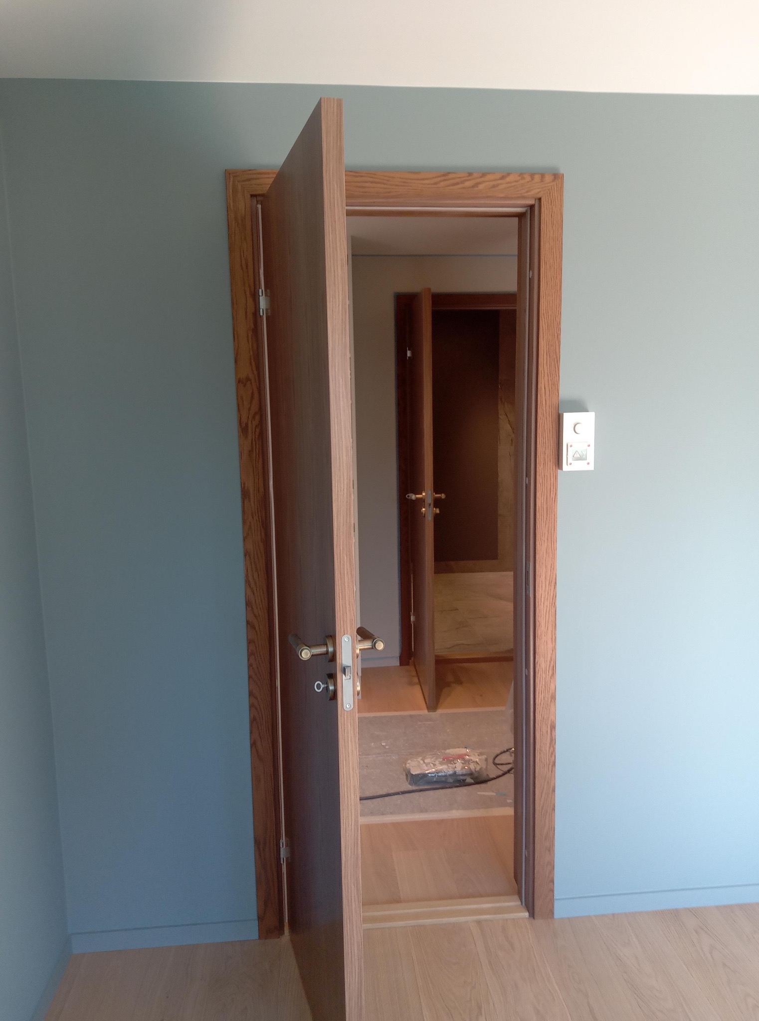 Montaż drewnianych drzwi wewnętrznych z ościeżnicą w pomieszczeniu o błękitnych ścianach i jasnej podłodze. Widoczne narzędzia i kolejne drzwi w głębi.