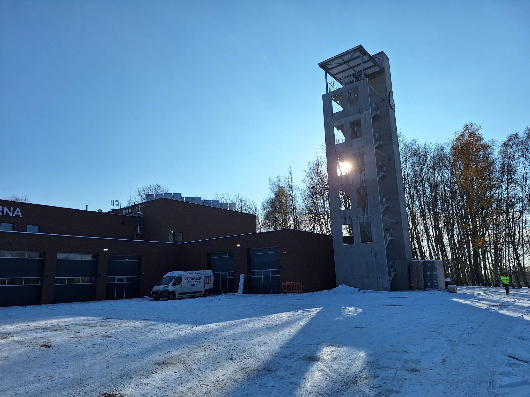 Nowoczesny budynek straży pożarnej z wieżą treningową w surowym stanie, oświetlony słońcem, zimowy krajobraz z widocznym vanem firmy budowlanej.