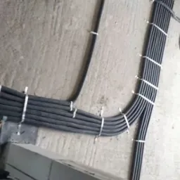 Równo ułożone, czarne kable w peszlach, przymocowane do szarej ściany za pomocą białych opasek zaciskowych. Widoczne przewody wychodzące ze ściany.