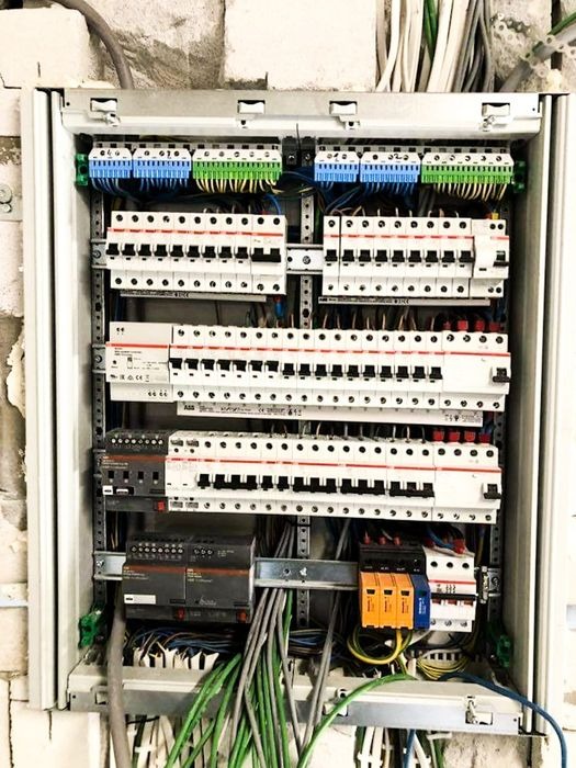 Otwarta skrzynka elektryczna z bezpiecznikami, okablowaniem i listwami zaciskowymi na tle surowej ściany z cegieł. Widok z bliska na elementy instalacji.