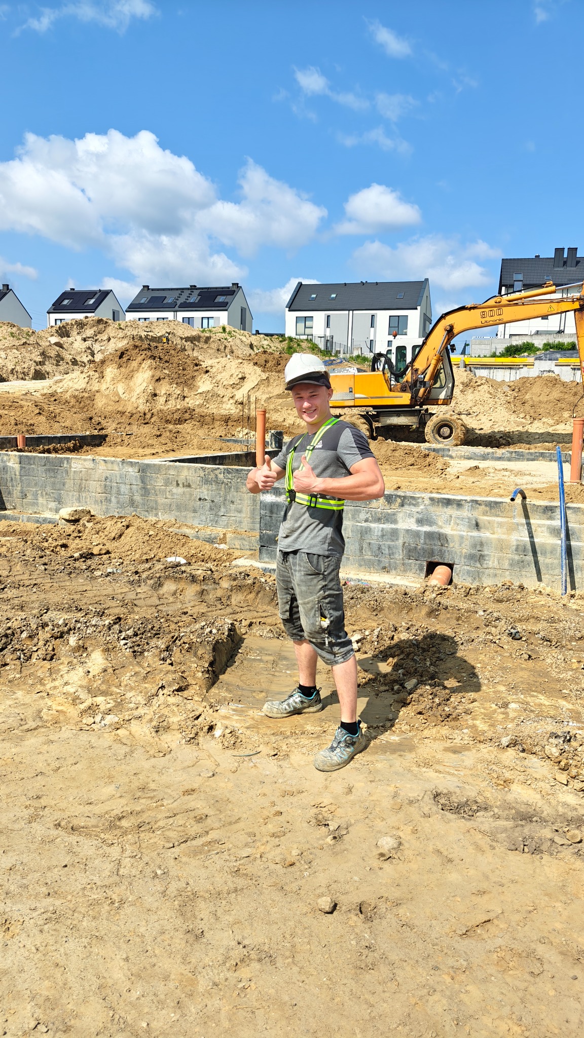 Uśmiechnięty pracownik budowlany w kasku i kamizelce odblaskowej, na tle wykopu pod fundamenty i koparki. Domy w tle.