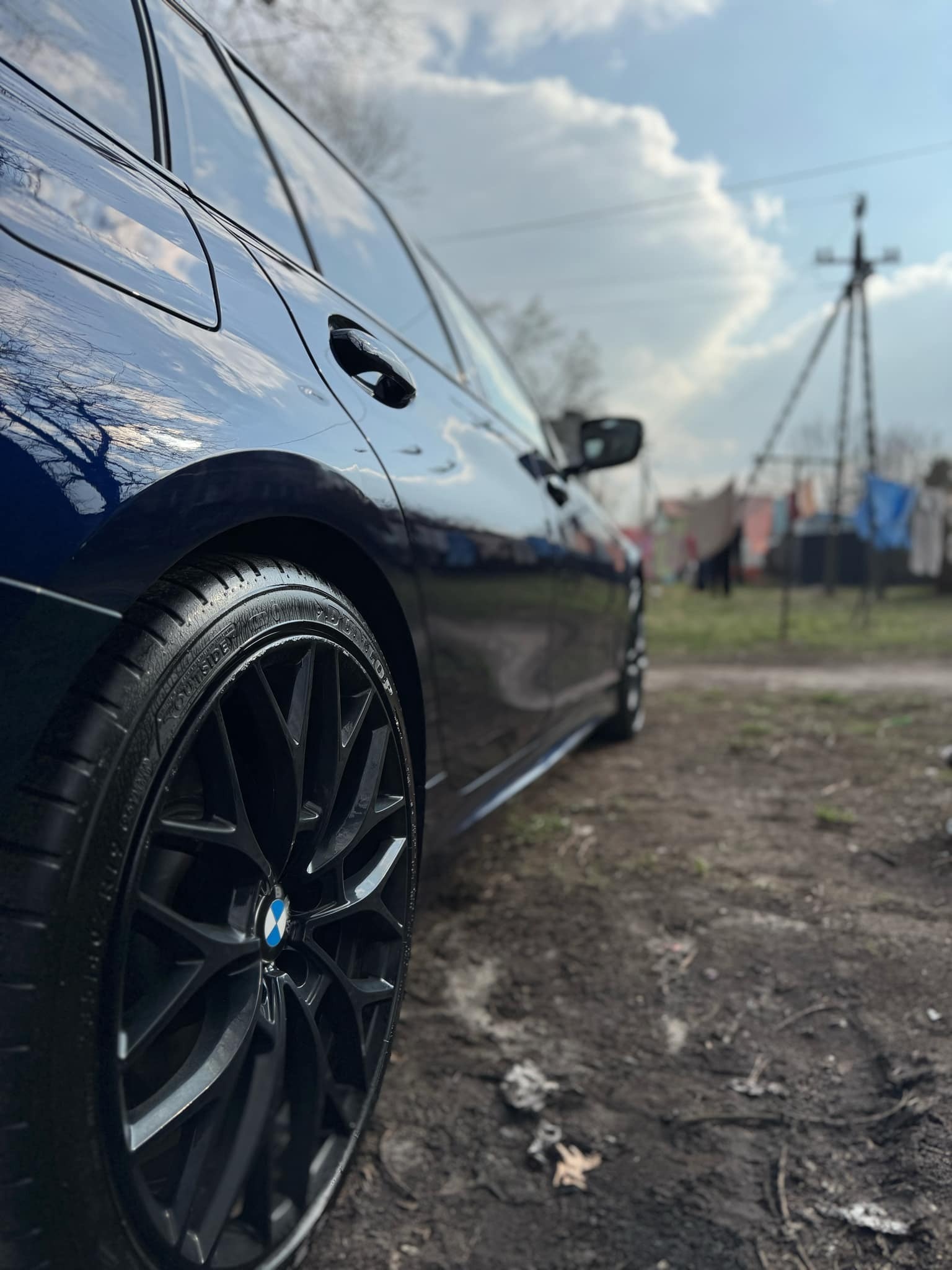 Błyszczący, granatowy samochód BMW po umyciu, z bliska widoczne czyste felgi na tle pochmurnego nieba i wiejskiego krajobrazu.