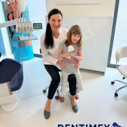GABINET STOMATOLOGICZNY ''DENTIMEX'' Edyta Mazur - Uśmiechnięta dentystka z pacjentką trzymającą aparat ortodontyczny w gabinecie, na tle sprzęt stomatologiczny i logo firmy Dentimex.