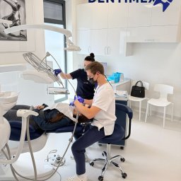 GABINET STOMATOLOGICZNY ''DENTIMEX'' Edyta Mazur - Nowoczesny gabinet stomatologiczny Dentimex: dentysta w masce i asystentka podczas zabiegu na pacjencie leżącym na niebieskim fotelu. Minimalistyczny wystrój, białe meble.