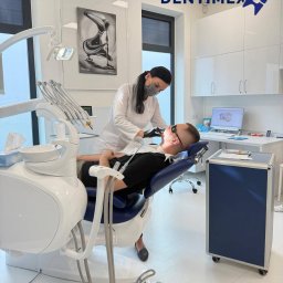 GABINET STOMATOLOGICZNY ''DENTIMEX'' Edyta Mazur - Stomatolog w masce podczas zabiegu u pacjenta leżącego na fotelu dentystycznym. W tle nowoczesny gabinet z obrazem i sprzętem.