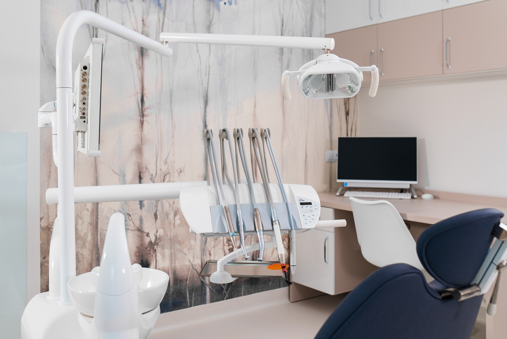 Nowoczesny gabinet stomatologiczny z białym fotelem, narzędziami i lampą. W tle biurko z komputerem i szafki. Tapeta w delikatne wzory drzew dodaje wnętrzu charakteru.