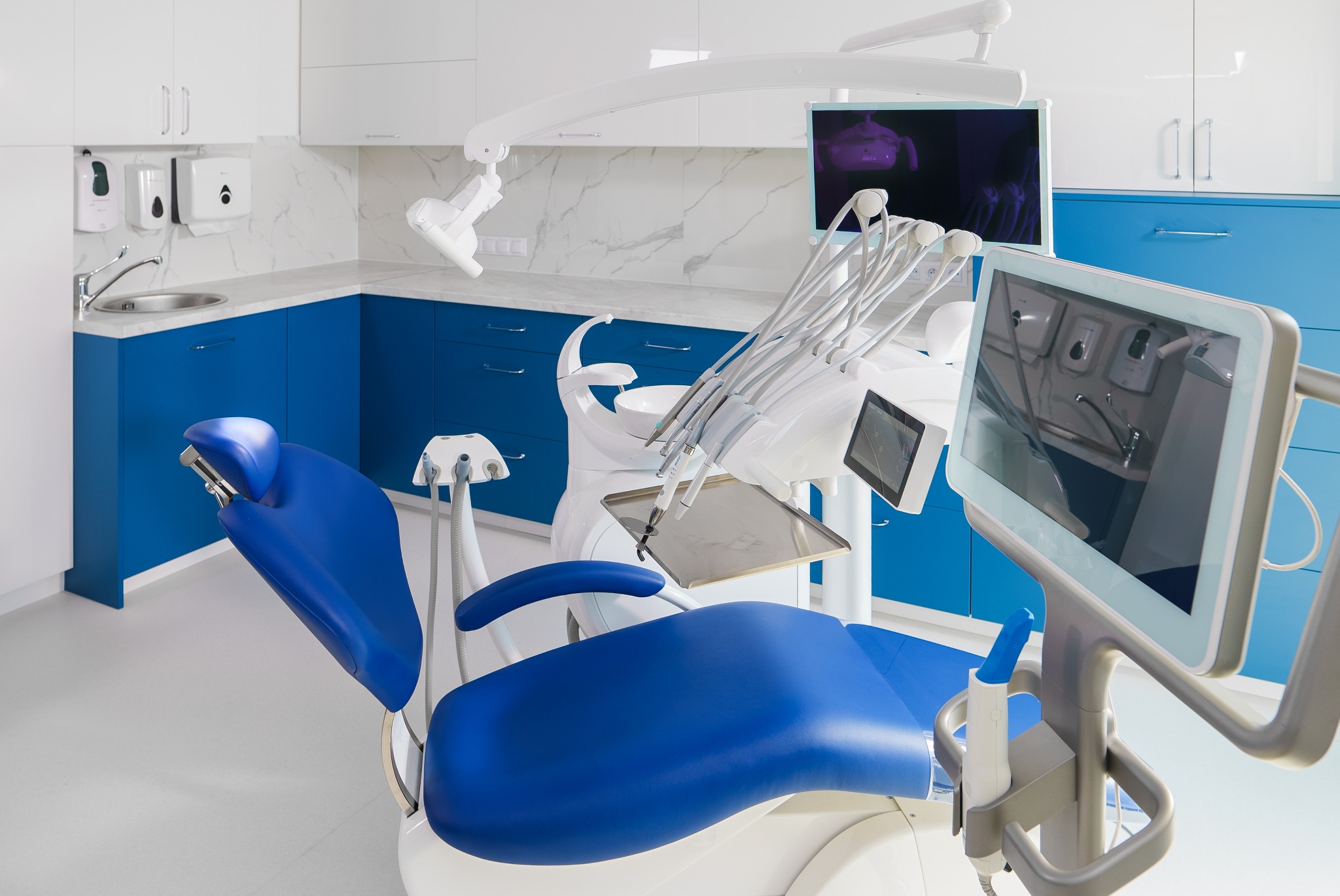 Nowoczesny gabinet stomatologiczny z niebieskim fotelem, białymi szafkami i marmurowym blatem. Widoczne narzędzia i monitory.