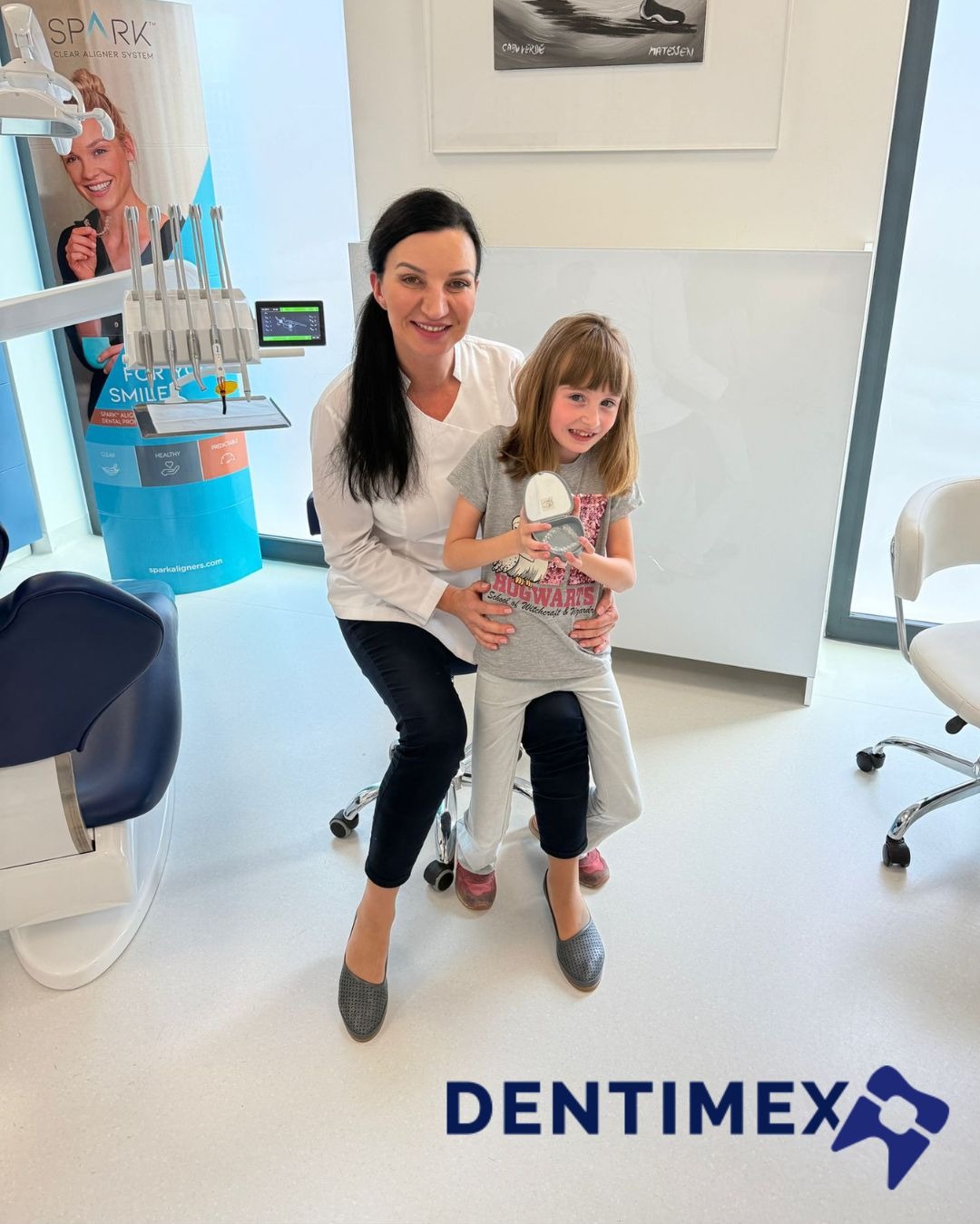 Uśmiechnięta dentystka z pacjentką trzymającą aparat ortodontyczny w gabinecie, na tle sprzęt stomatologiczny i logo firmy Dentimex.