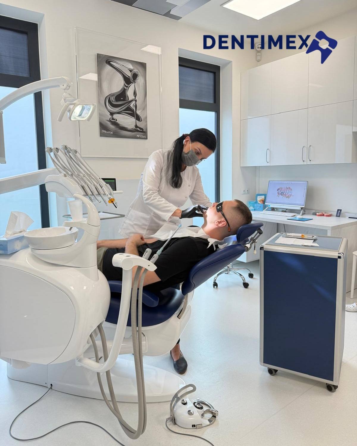 Stomatolog w masce podczas zabiegu u pacjenta leżącego na fotelu dentystycznym. W tle nowoczesny gabinet z obrazem i sprzętem.