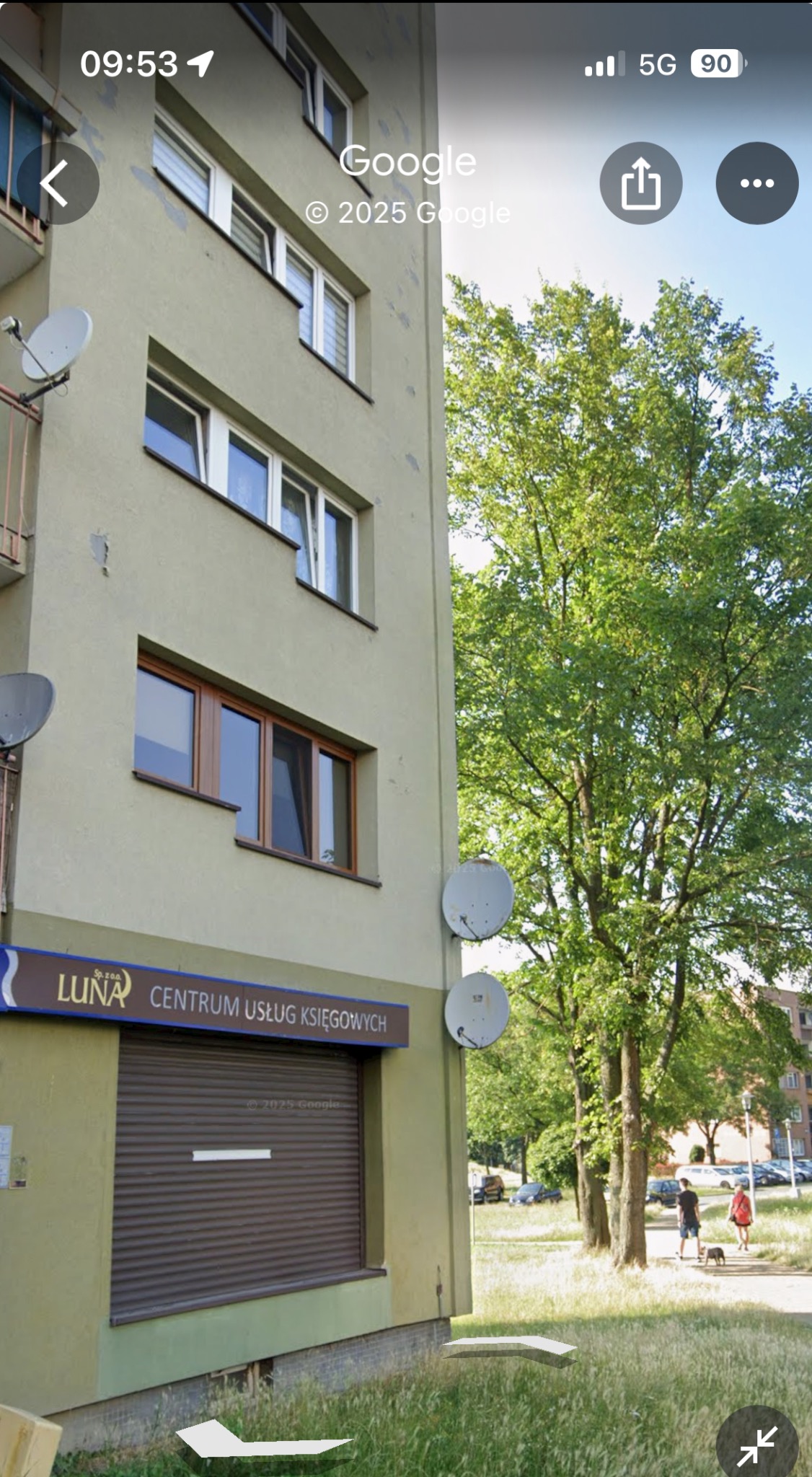 Budynek z szyldem 'LUNA Centrum Usług Księgowych' i zamkniętą roletą. Widoczne okna, anteny satelitarne i drzewa w tle. Ujęcie z dołu, w słoneczny dzień.