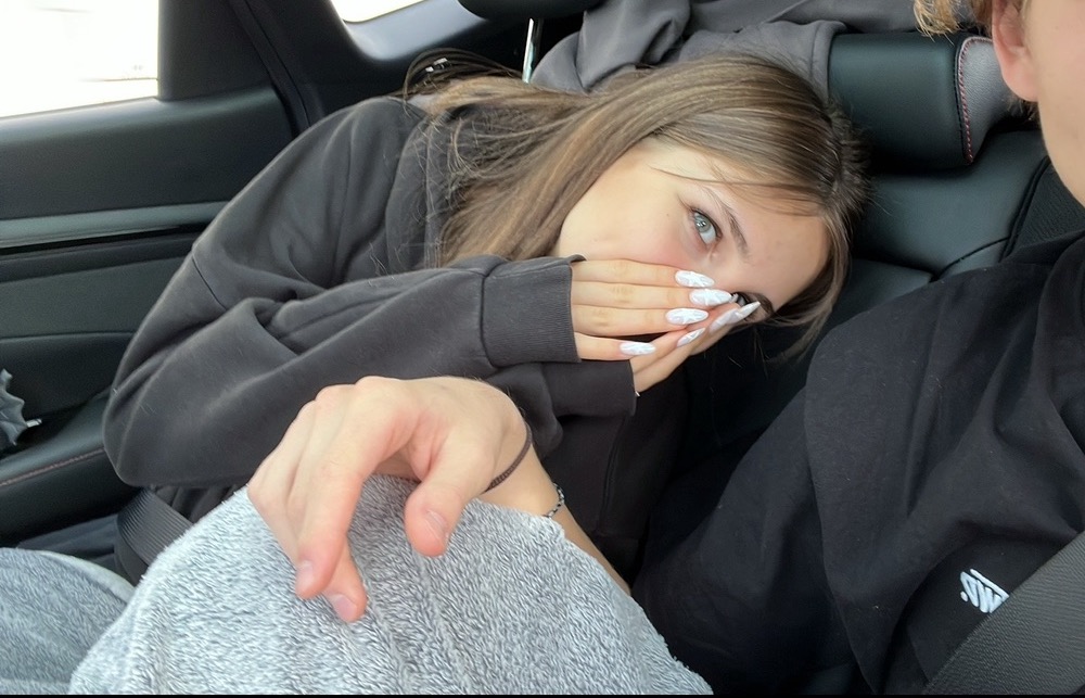 Nastolatka z długimi, wzorzystymi paznokciami zakrywa usta w samochodzie, obok siedzi druga osoba z zapiętym pasem bezpieczeństwa.