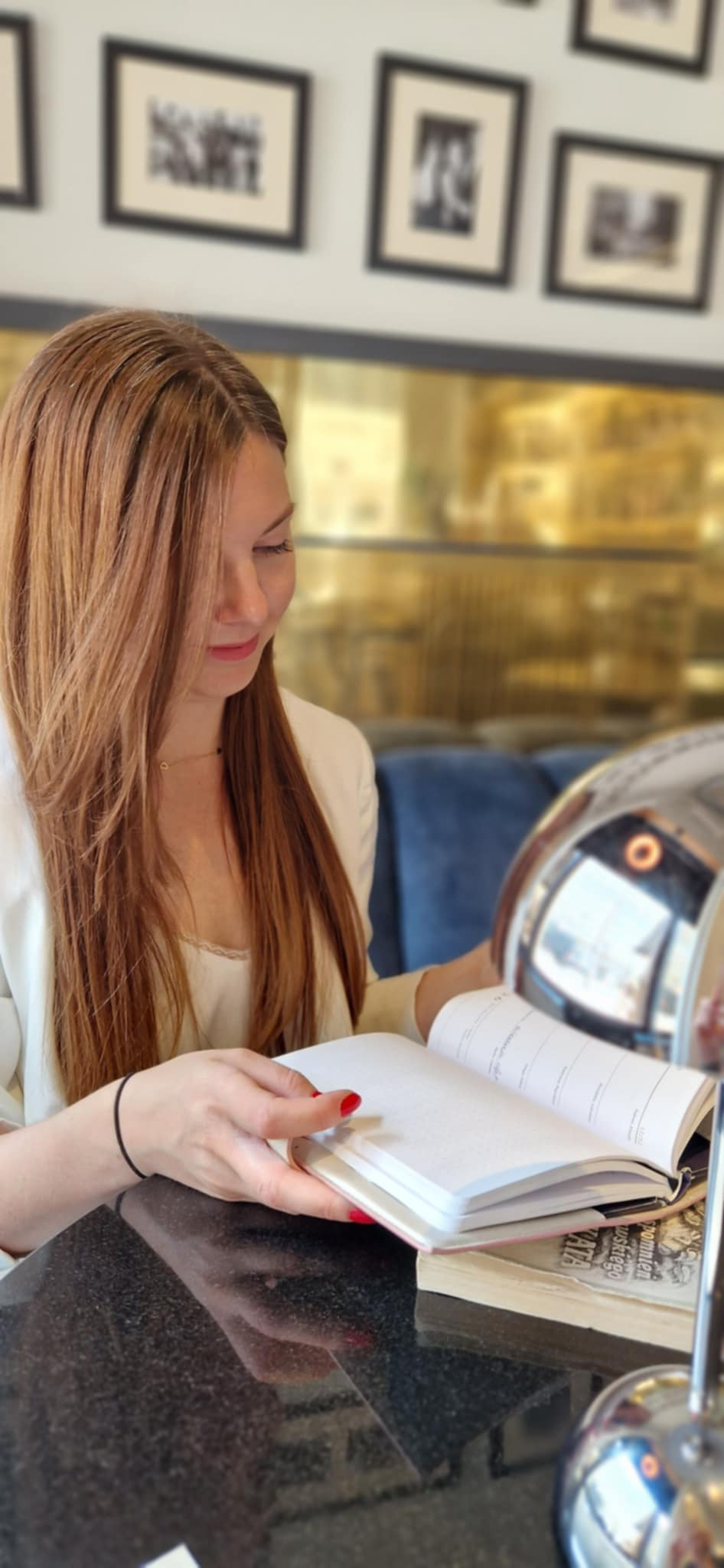 Kobieta z rudymi włosami przegląda notatnik przy stoliku w kawiarni. W tle widoczne dekoracje ścienne i złote elementy wystroju.