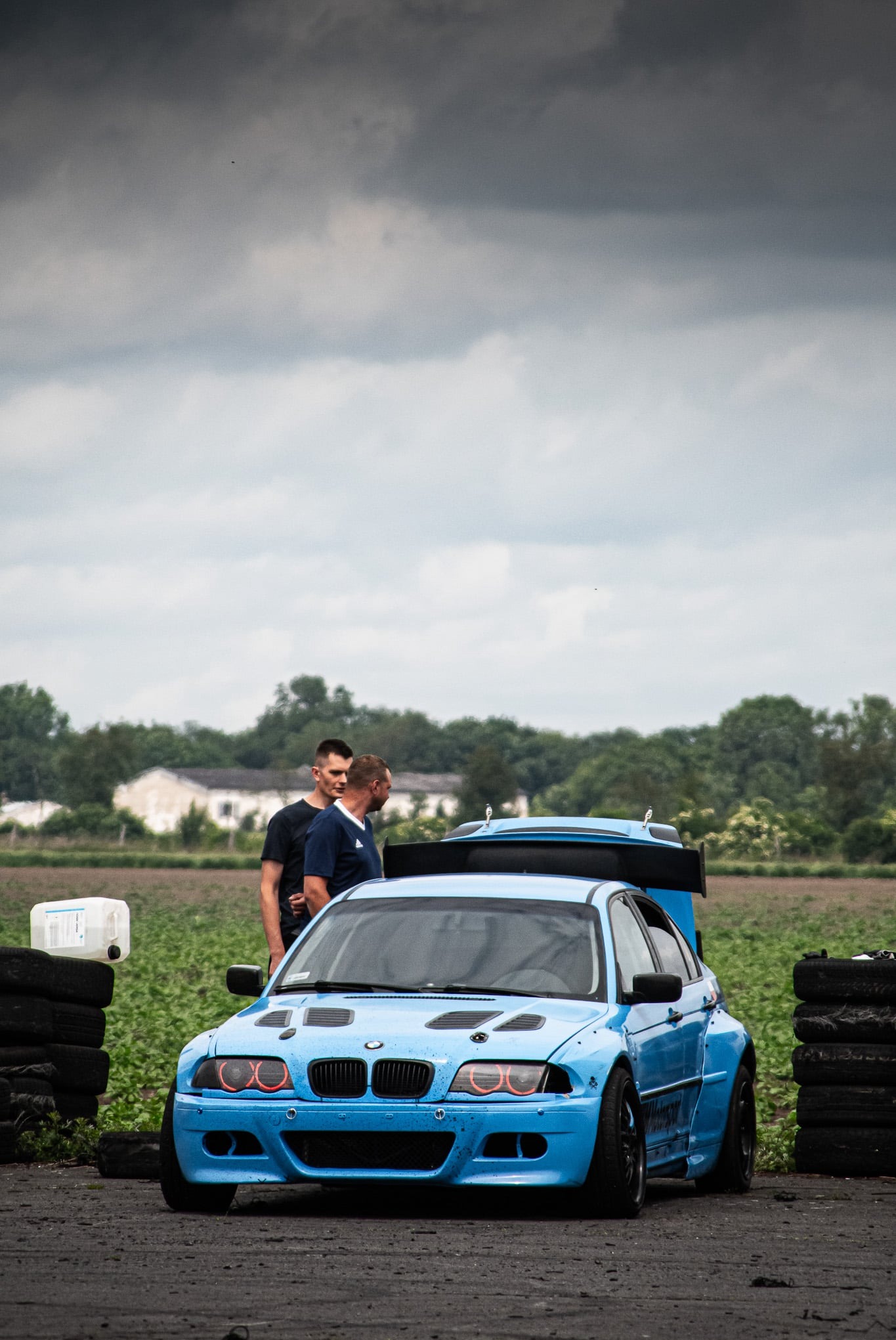 Błękitne BMW E46 z body kit na tle pola, dwóch mężczyzn i sterty opon. Tuningowany samochód sportowy, widok z przodu, pochmurne niebo w tle. Detale karoserii.