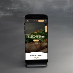 Aleksandra Godlewska Papaja Studio - Smartfon prezentuje stronę internetową pizzerii Pod Kłosem z Suwałk, z opcją zamówienia online i menu. Widoczne logo, adres i ikony mediów społecznościowych.
