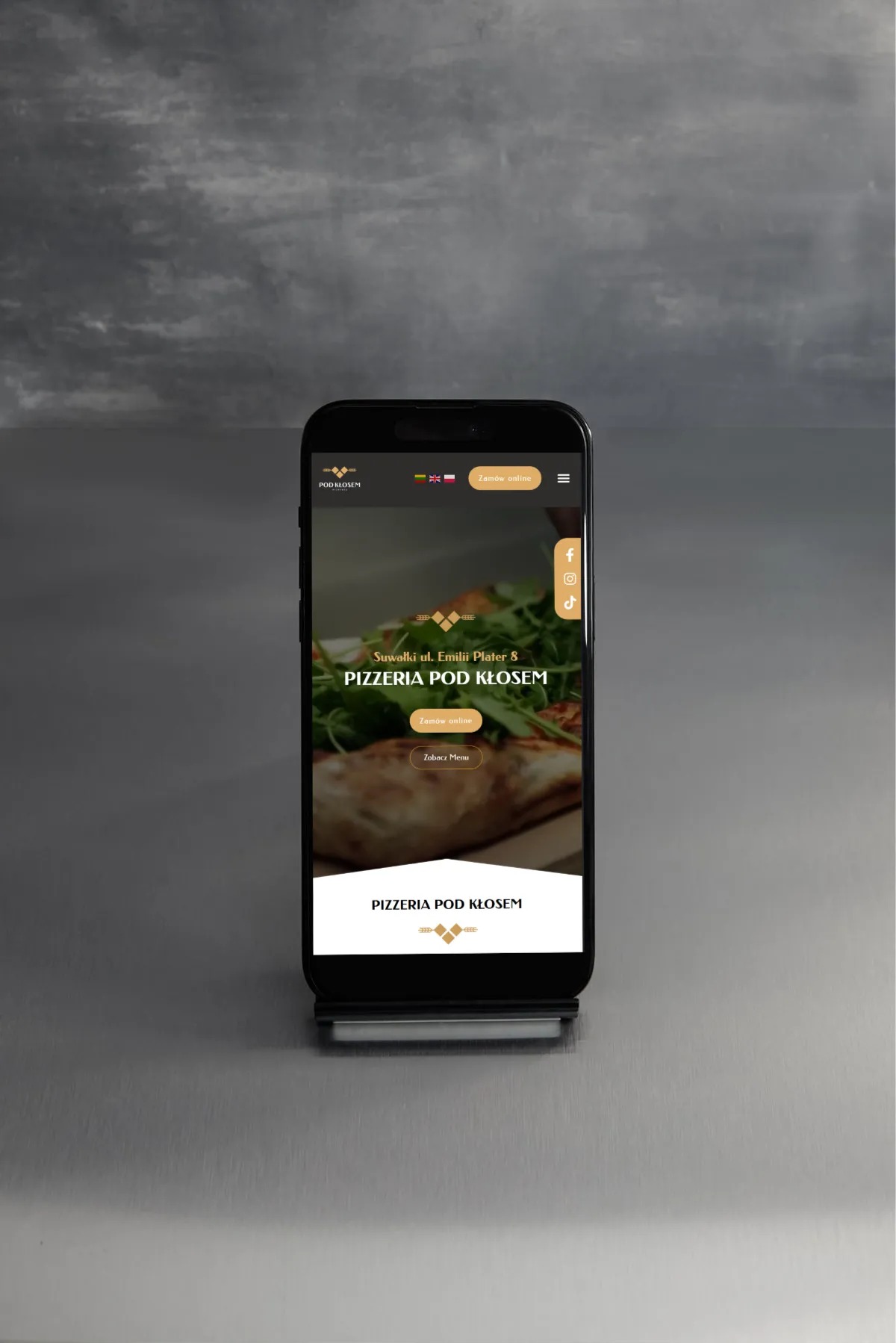Smartfon prezentuje stronę internetową pizzerii Pod Kłosem z Suwałk, z opcją zamówienia online i menu. Widoczne logo, adres i ikony mediów społecznościowych.