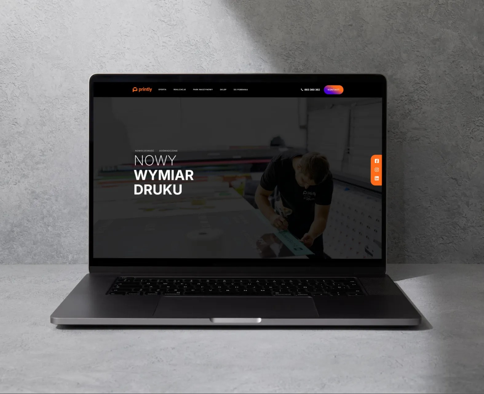 Laptop prezentuje stronę internetową drukarni Printly z hasłem 'Nowy Wymiar Druku' i zdjęciem pracownika przy pracy. Widoczne logo, menu i ikony social media.