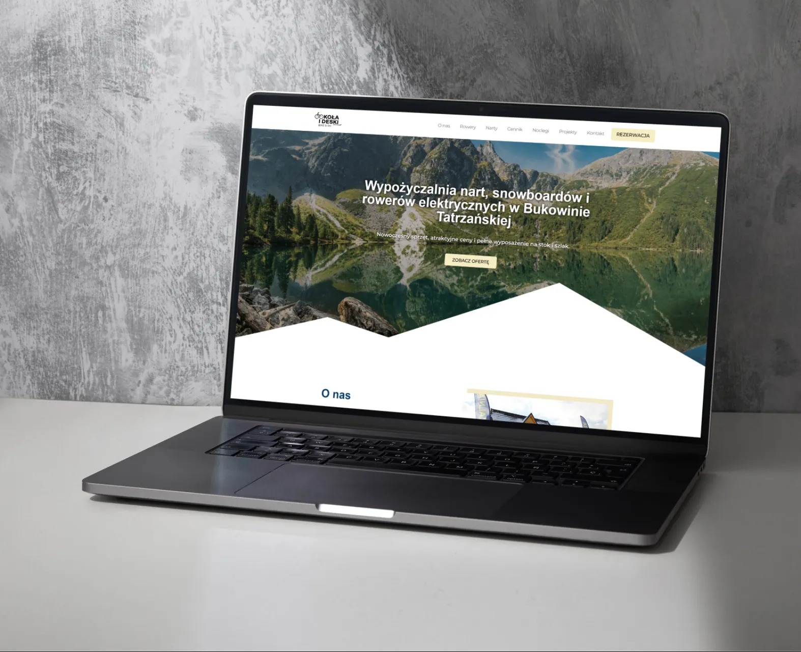 Laptop prezentuje stronę wypożyczalni sprzętu zimowego i rowerów elektrycznych. Widok na jezioro w górach. Szara obudowa laptopa na jasnym tle.