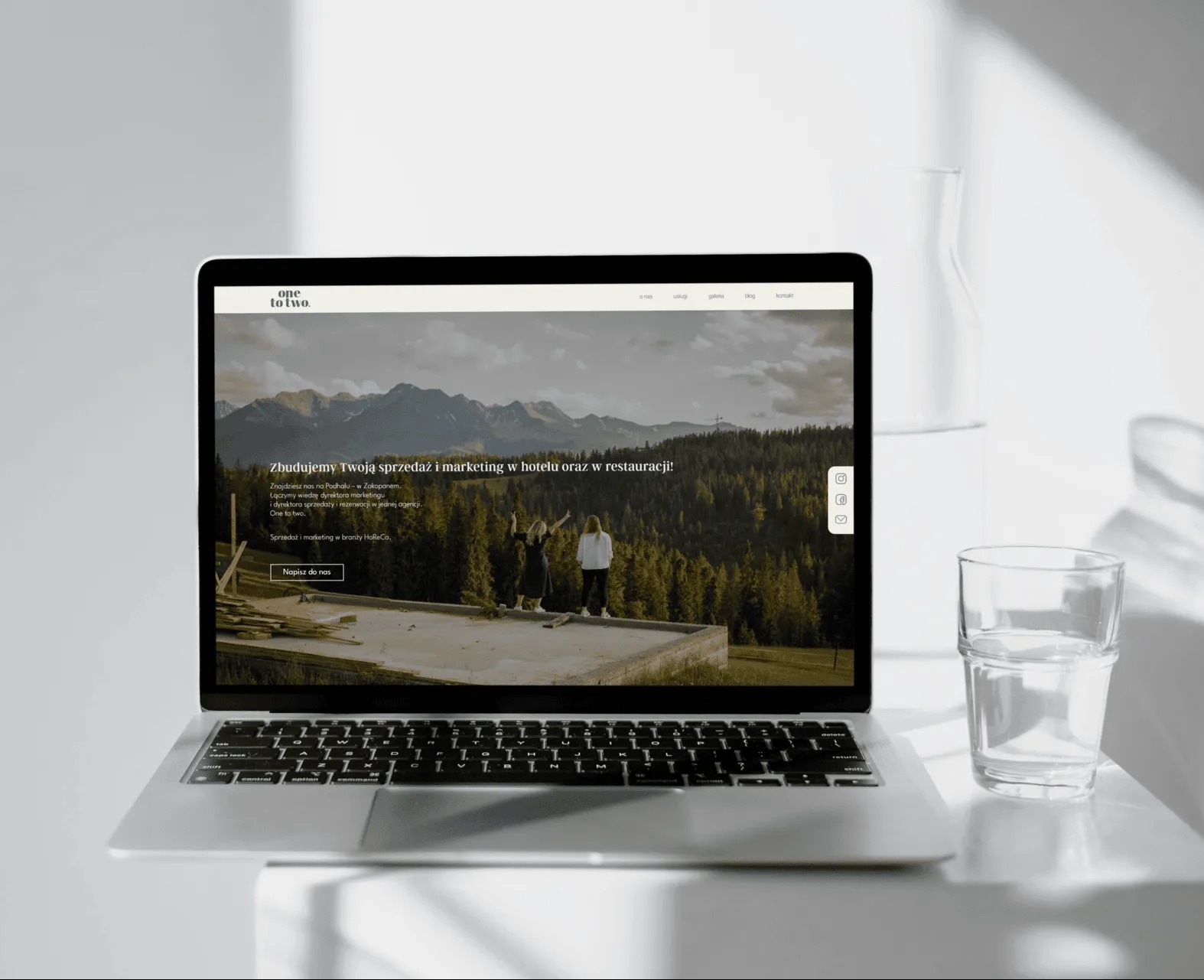 Laptop wyświetla stronę internetową 'one to two' z hasłem: Zbudujemy Twoją sprzedaż i marketing w hotelu oraz restauracji!. W tle góry, obok szklanka wody.