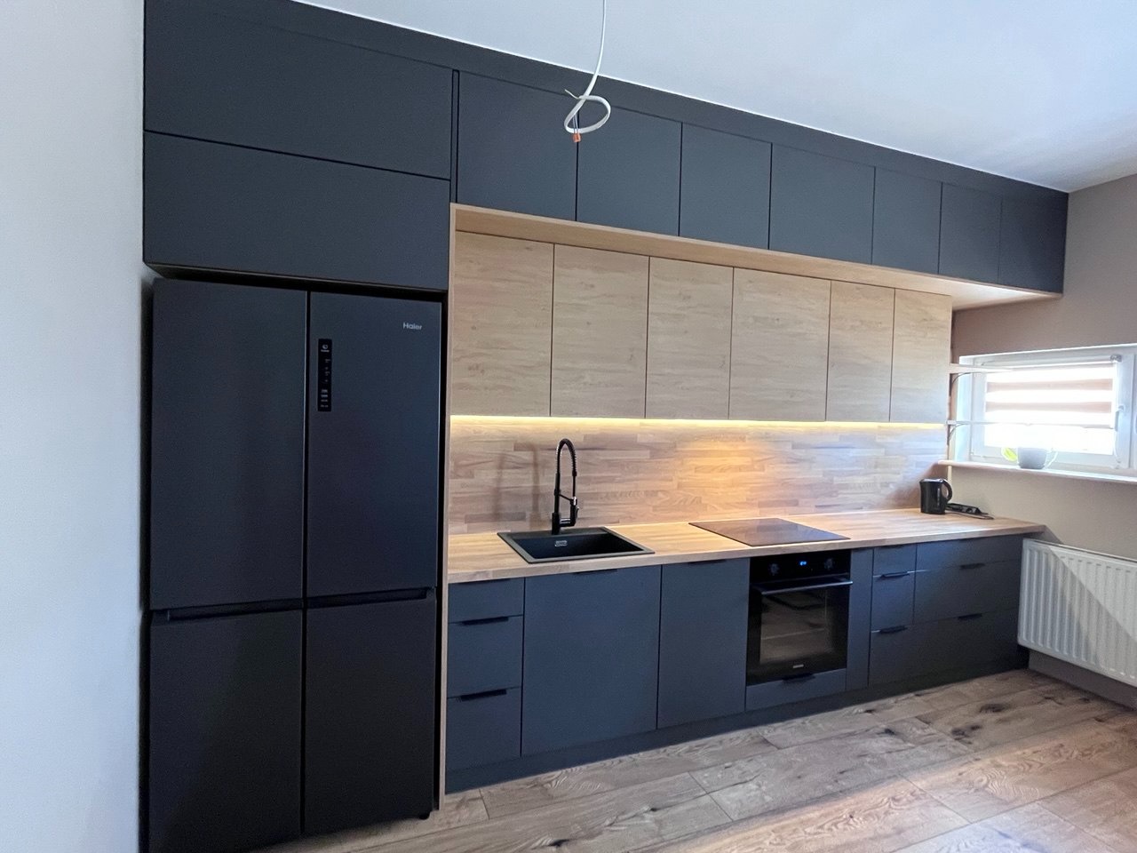 Nowoczesna kuchnia z ciemnymi szafkami, drewnianym blatem i dużą, czarną lodówką. Górne szafki w dwóch kolorach. Minimalistyczny design, oświetlenie LED pod szafkami.