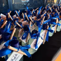Rząd identycznych, upominkowych buteleczek z płynem, zdobionych niebieską wstążką i etykietą Bluebell Event Agency. Drewniane patyczki wystają z korków.