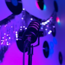 Oleksandr Panait Bluebell Agency - Mikrofon PodMic na statywie w fioletowym oświetleniu, tło z dekoracjami przypominającymi płyty winylowe. Sprzęt nagłośnieniowy w studio.