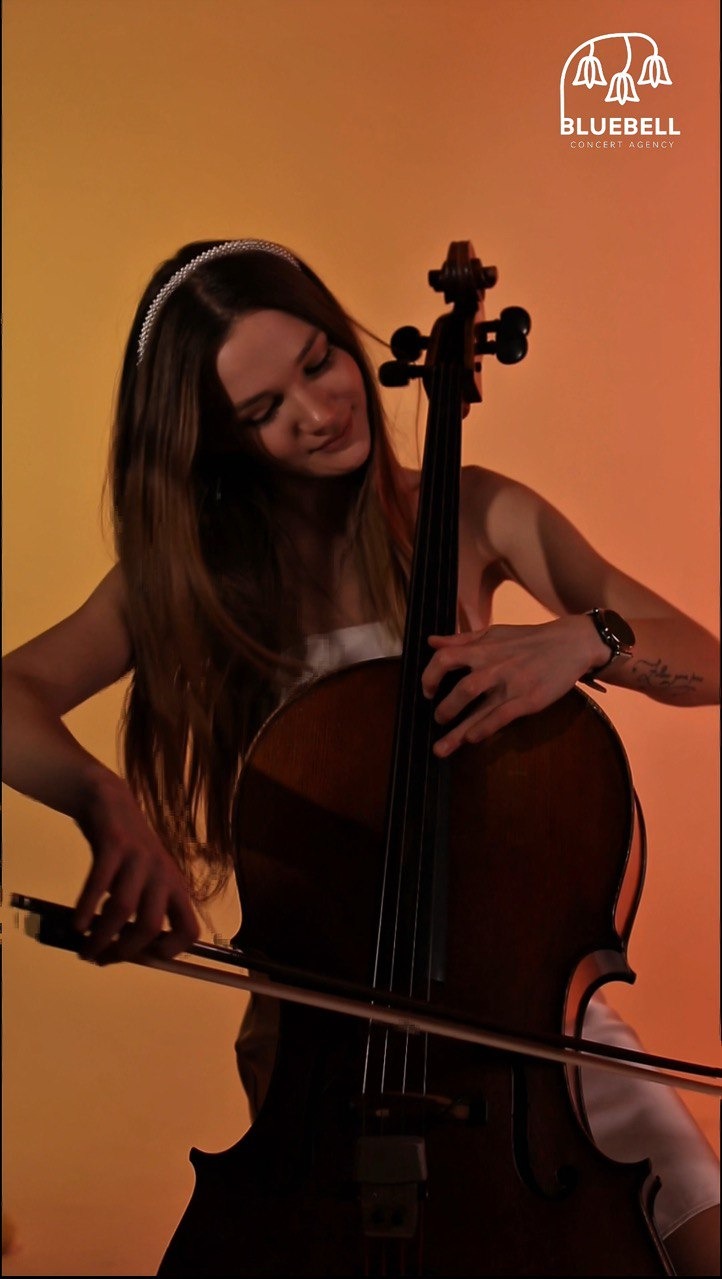 Młoda kobieta z opaską gra na wiolonczeli na pomarańczowym tle, widoczny fragment logo Bluebell Concert Agency w prawym górnym rogu.
