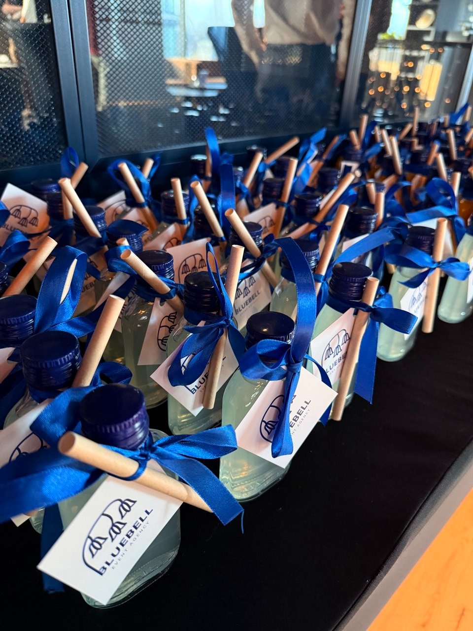Rząd identycznych, upominkowych buteleczek z płynem, zdobionych niebieską wstążką i etykietą Bluebell Event Agency. Drewniane patyczki wystają z korków.