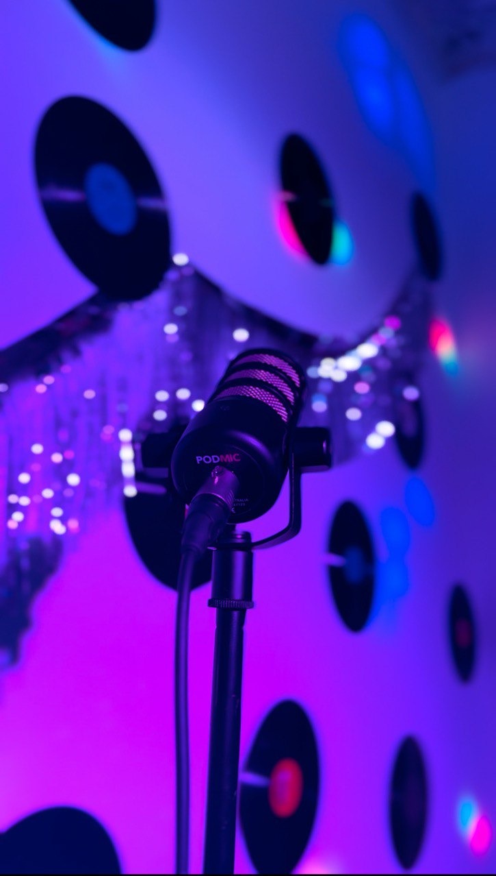 Mikrofon PodMic na statywie w fioletowym oświetleniu, tło z dekoracjami przypominającymi płyty winylowe. Sprzęt nagłośnieniowy w studio.