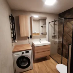 Nowoczesna łazienka w beżach i brązach z drewnianymi akcentami: kabina prysznicowa typu walk-in z czarną ramą, umywalka z szafką, pralka pod drewnianym blatem i dekoracyjna ściana z geometrycznym...