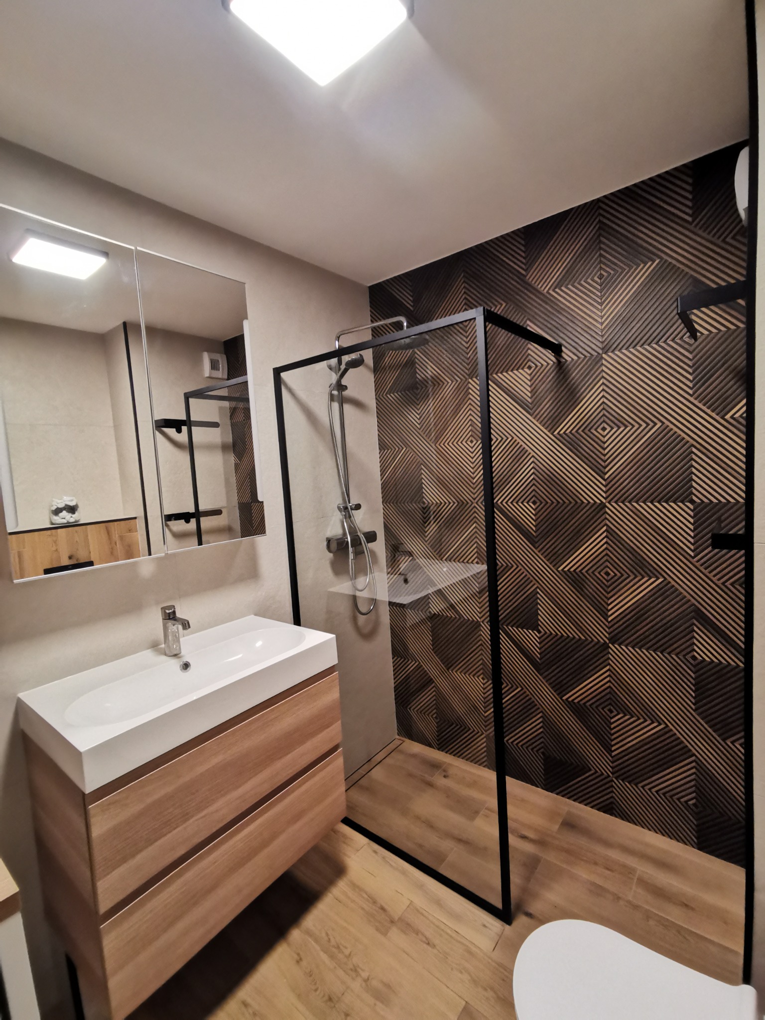 Nowoczesna łazienka z kabiną prysznicową typu walk-in z czarnymi profilami, umywalką nablatową na drewnianej szafce i ścianą wyłożoną dekoracyjnymi płytkami w geometryczny wzór.