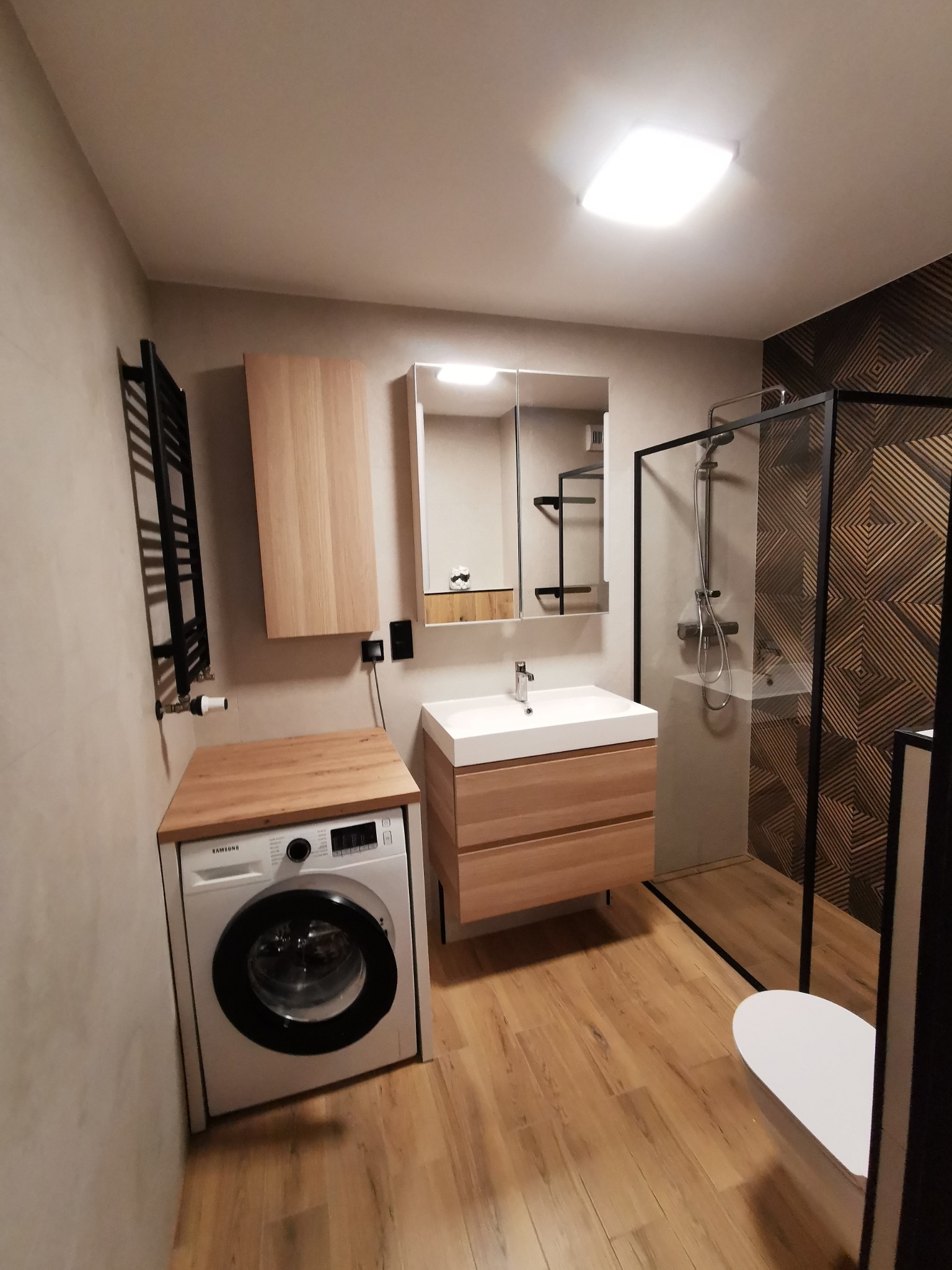 Nowoczesna łazienka w beżach i brązach z drewnianymi akcentami: kabina prysznicowa typu walk-in z czarną ramą, umywalka z szafką, pralka pod drewnianym blatem i dekoracyjna ściana z geometrycznym...
