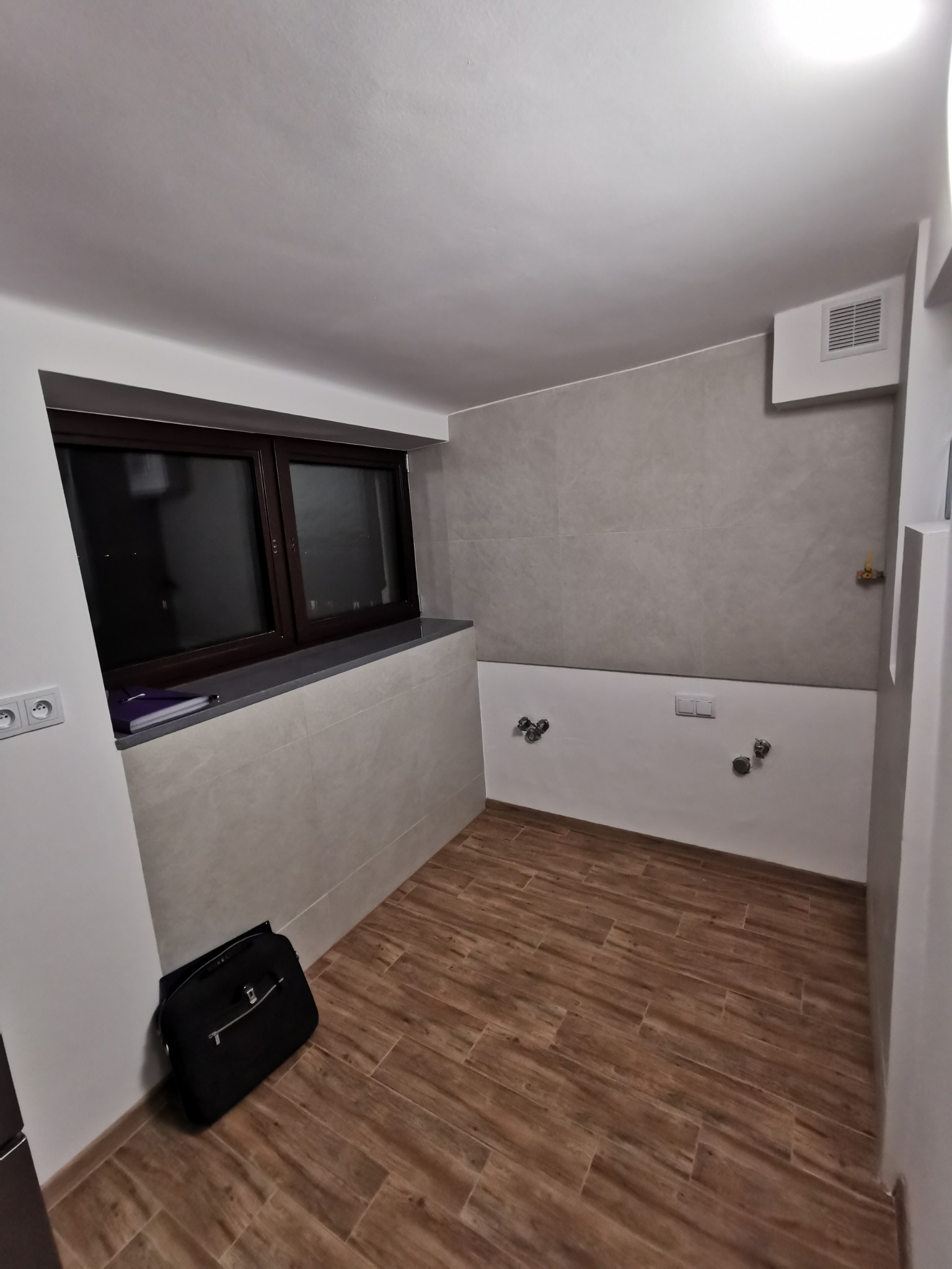 Przygotowana do montażu armatury łazienka z wykończonymi ścianami w szarej glazurze i drewnopodobną podłogą, widoczne wystające przyłącza wodne i kanalizacyjne oraz czarna torba robocza przy ścianie.