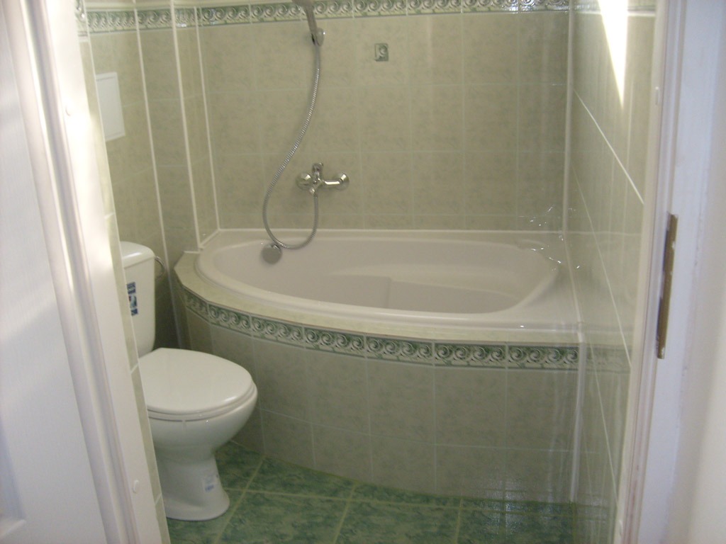 Jasna łazienka z narożną wanną obudowaną płytkami z dekoracyjnym wzorem, toaleta kompaktowa z widoczną naklejką producenta, całość utrzymana w odcieniach zieleni i bieli.