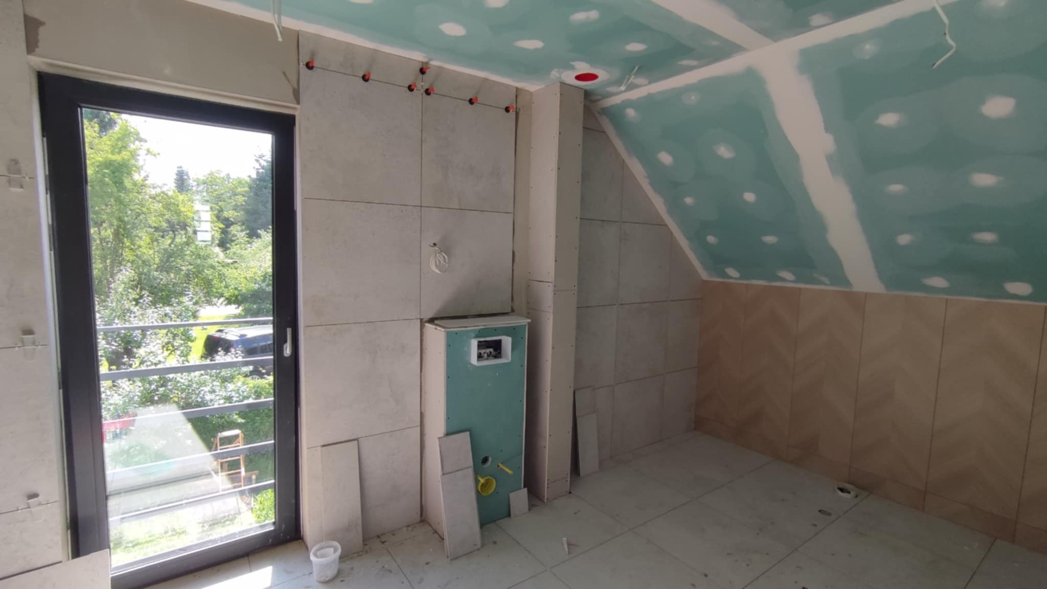 Łazienka na poddaszu w trakcie wykańczania: położone płytki, zamontowana stelaż podtynkowy, sufit z płyt karton-gips. Okno balkonowe z widokiem na zieleń.