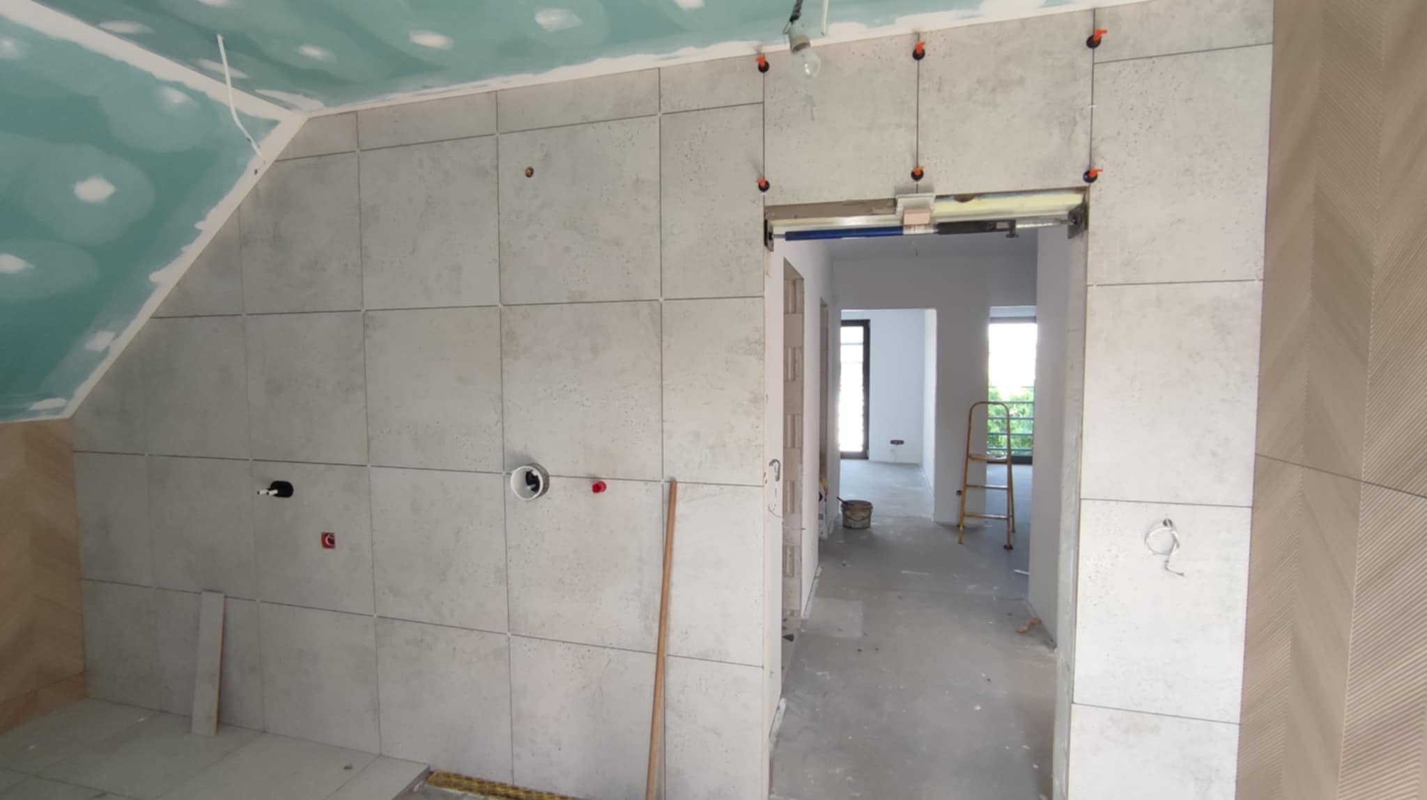 Wnętrze w trakcie wykańczania: ułożone płytki na ścianach, widoczne otwory instalacyjne, sufit z płyt karton-gips, drabina w tle.