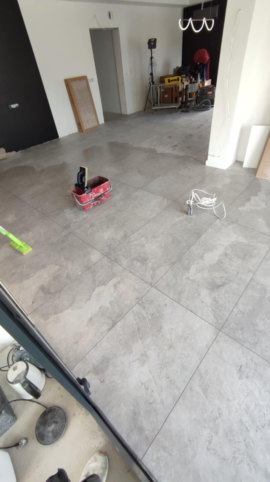 Układanie szarych płytek podłogowych w dużym formacie. Widoczne narzędzia glazurnicze: wiadro, mop, poziomica. W tle sprzęt budowlany i oświetlenie.