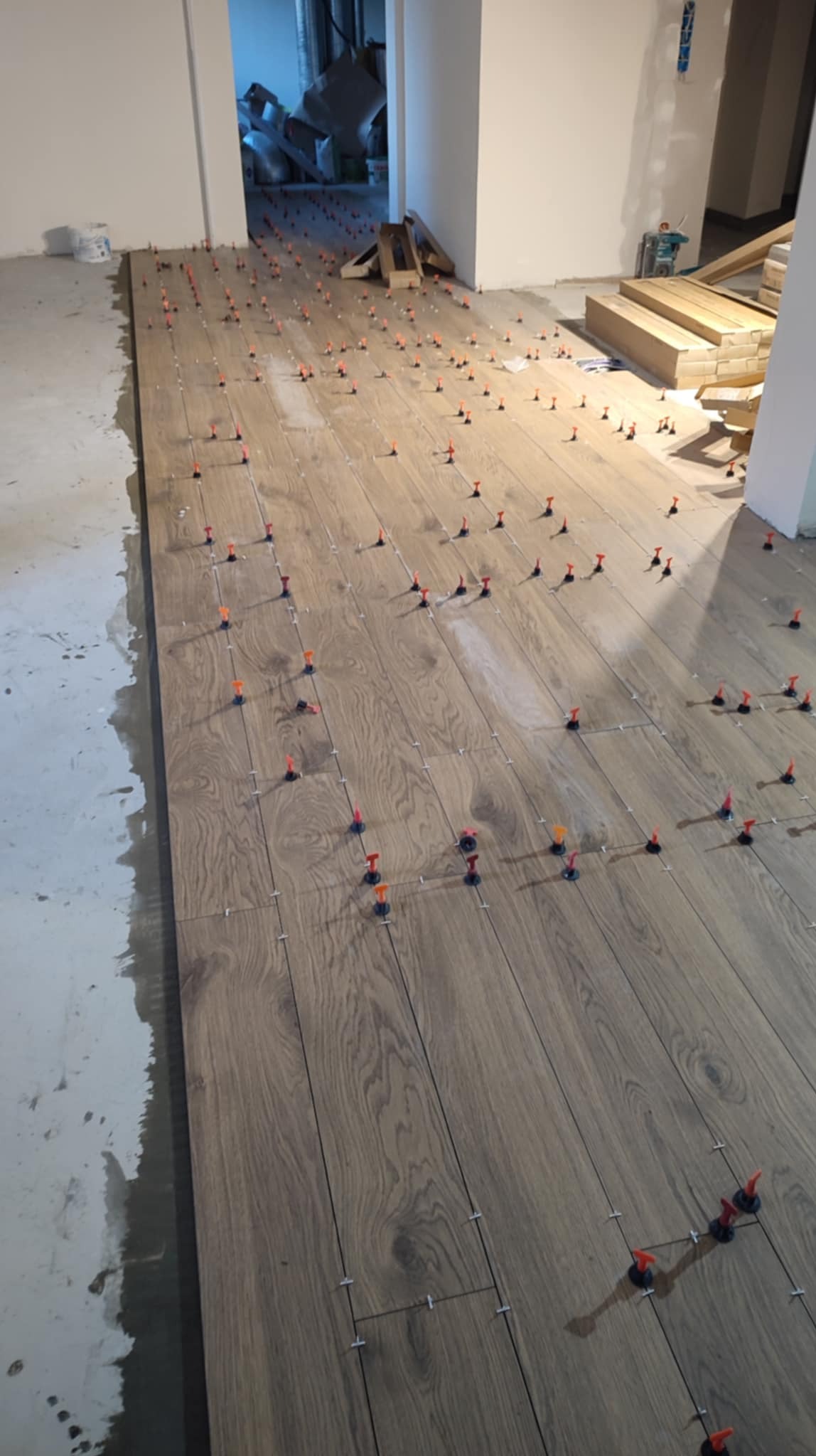 Układanie podłogi z paneli imitujących drewno z użyciem klipsów poziomujących. Widoczny fragment surowego podłoża i materiały budowlane w tle.