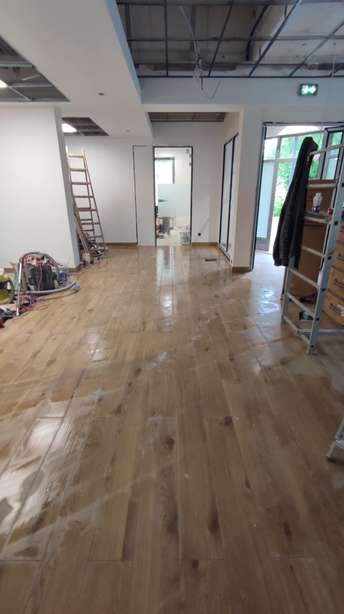 Wnętrze biura w trakcie remontu: świeżo położona drewniana podłoga, drabina, narzędzia malarskie. Widoczne niedokończone sufity podwieszane i surowe ściany.